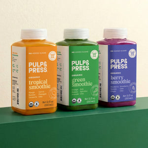 Three bottles of Pulp & Press smoothies in orange, green, and purple on a green surface with a beige background.