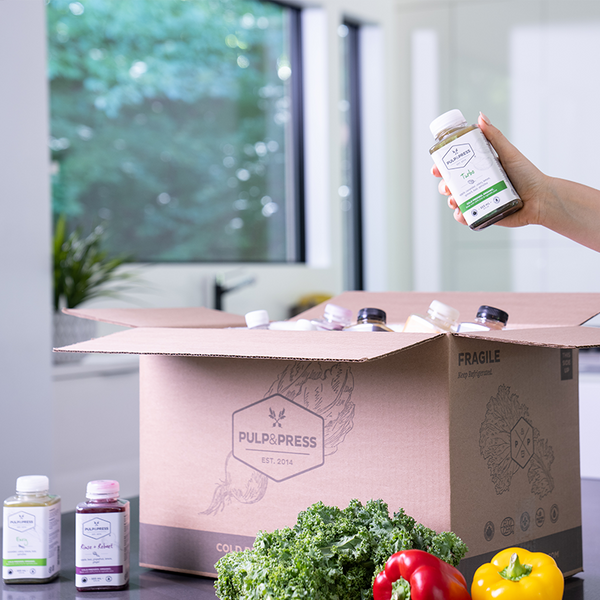 pulp and press cardboard shipping box on counter with juice cleanse and hand holing juice